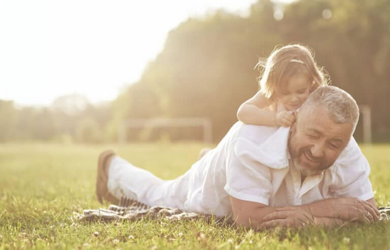 Young girl climbing on the back of her grandpa who's laying on the grass in the park