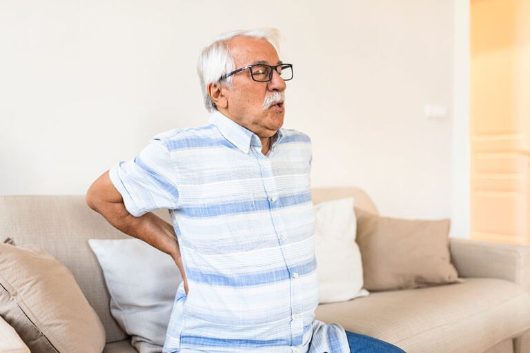 Older man exhaling slowly, holding his back while sitting on the edge of his couch